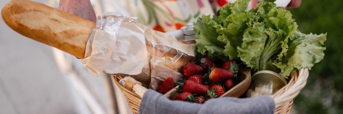 A woman in a floral dress holds a basket filled with bread, strawberries, and lettuce on her bicycle.